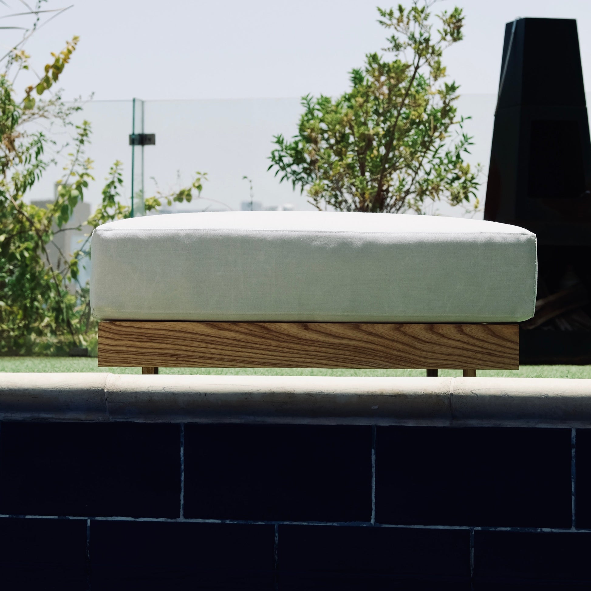 White outdoor ottoman with cushions by a poolside, surrounded by greenery.
