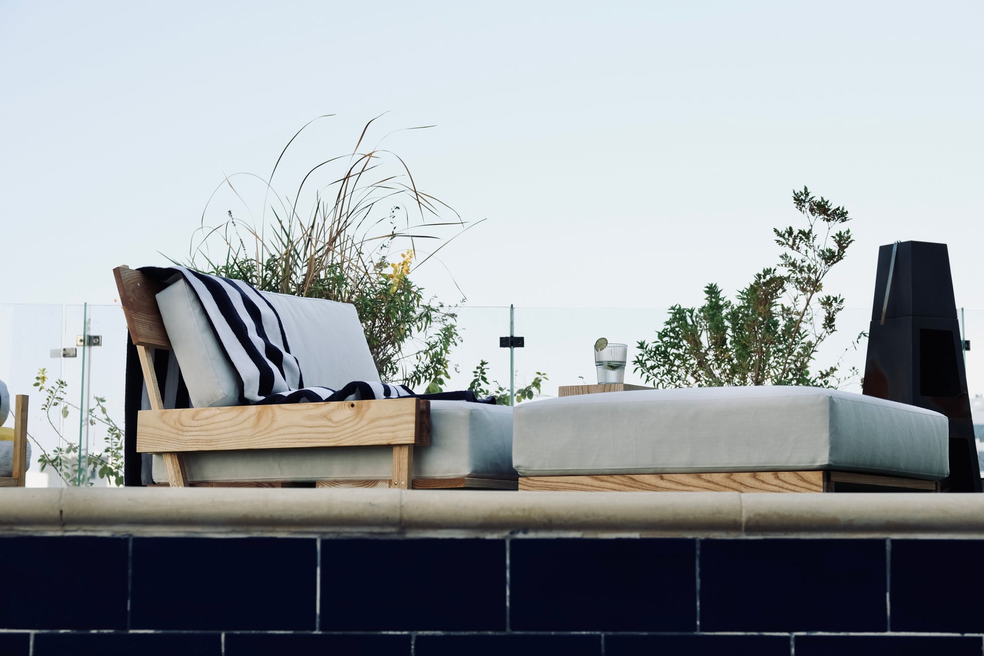 Outdoor furniture set with a chair and ottoman on a patio with plants and a clear sky.
